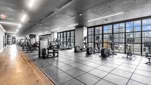 Uma academia espaçosa com diversos equipamentos de exercício, amplas janelas com vista para a cidade e piso de madeira.