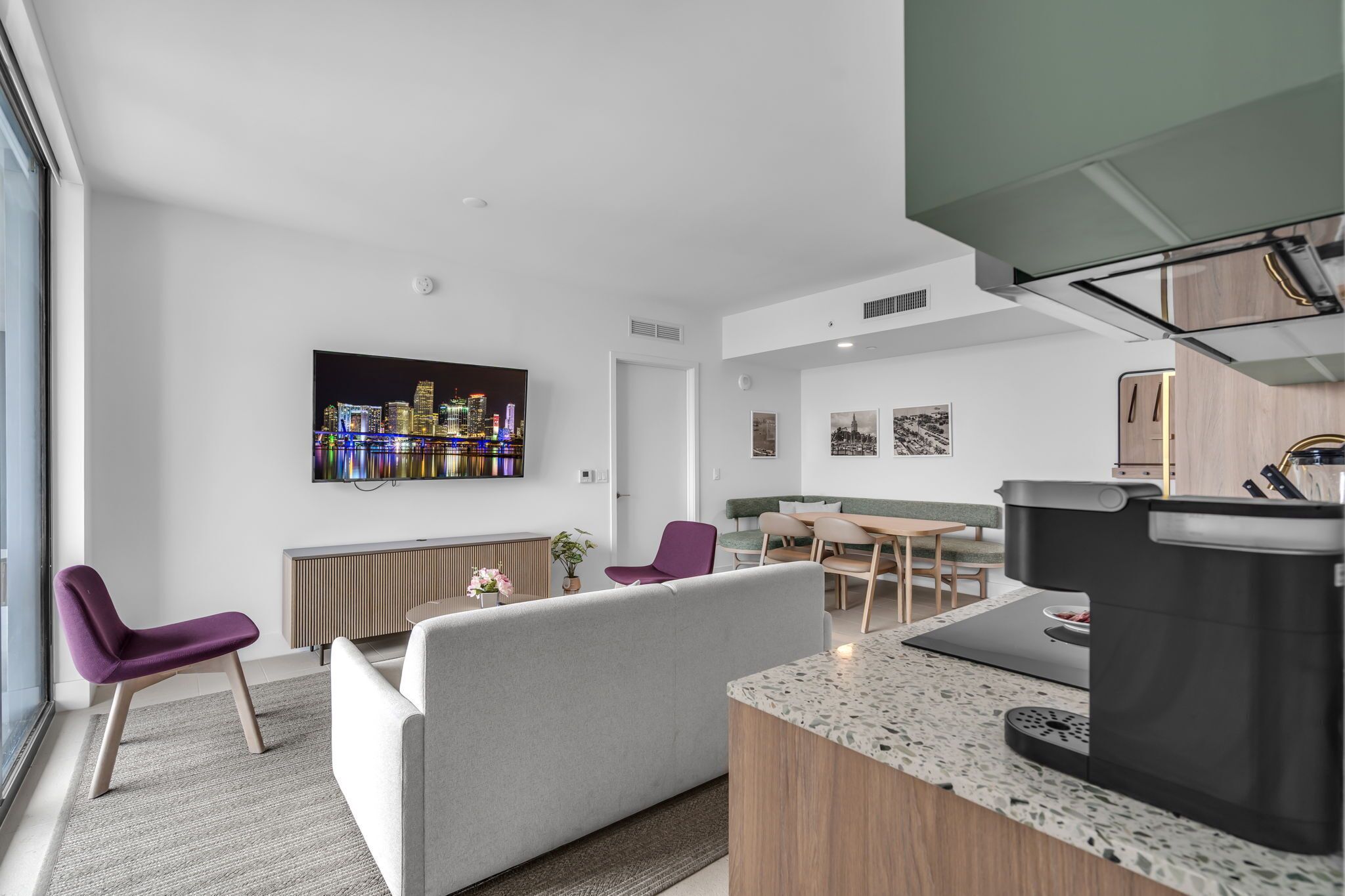 A modern living room with a sofa, chairs, a coffee machine, and a TV displaying a cityscape.