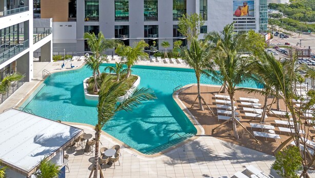 Uma área de piscina no terraço com palmeiras, espreguiçadeiras e vista para um prédio alto.