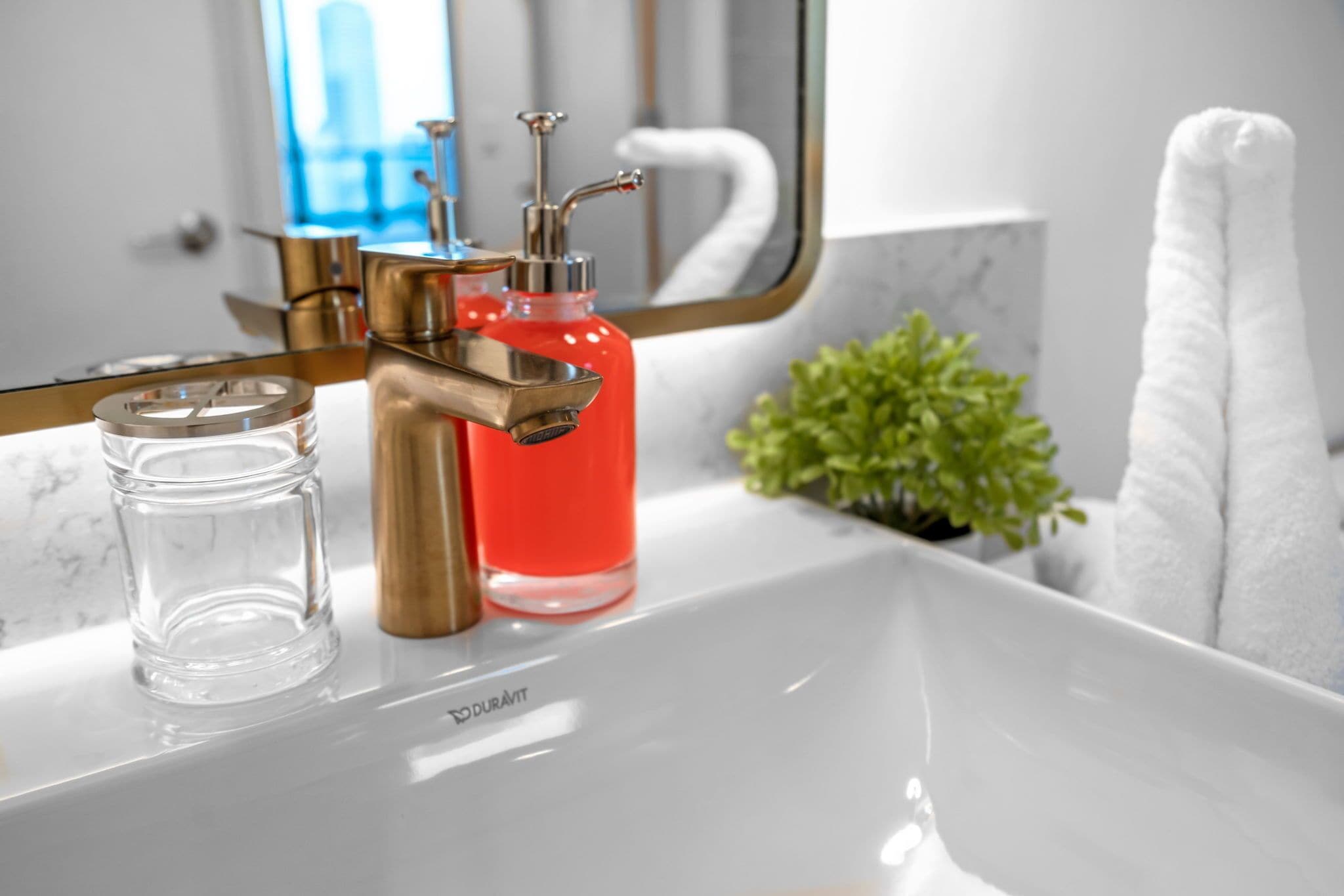 Un bagno con un lavandino bianco, un rubinetto dorato, una bottiglia rossa, un bicchiere trasparente e una pianta verde.