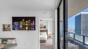 Quarto de hotel moderno com vista para a cidade, uma varanda com guarda-corpo, cama com cabeceira branca e área de estar com mesa e cadeiras.