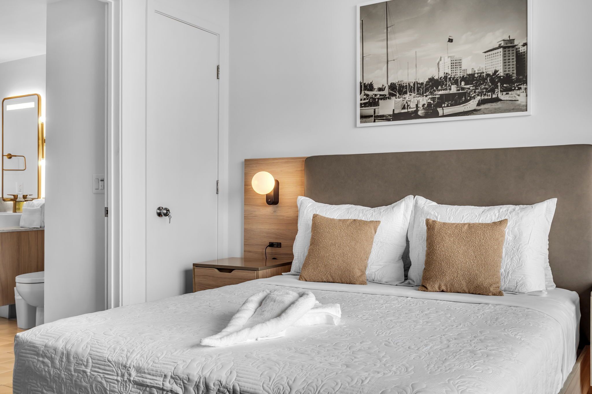 A neatly made bed with a white duvet, a brown pillow, and a white towel on top. There is a wooden headboard, a wall-mounted light, and a framed picture above the bed.