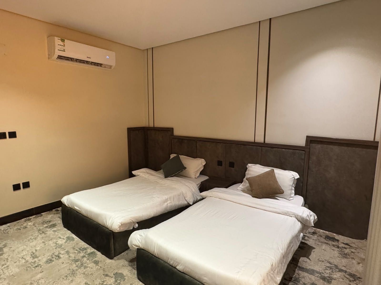 A hotel room with two single beds, a wall-mounted air conditioner, and a textured floor.