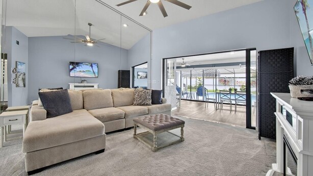 A modern living room with a sectional sofa, a glass coffee table, ceiling fans, and a large sliding glass door leading to an outdoor patio. - Footprints In The Sand | 3BR Private Pool Home | Near Palm Coast and Flagler Beaches (Palm Coast)
