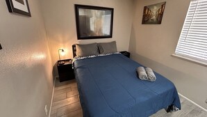 Une chambre à coucher comprenant un lit, une table de chevet, un miroir fixé au mur, une fenêtre avec des stores et un tableau accroché au mur.