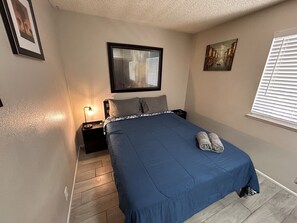 A bedroom with a bed, a nightstand, a wall-mounted mirror, a window with blinds, and a painting on the wall. - Jamming Jericho 2 bedrooms (Las Vegas)