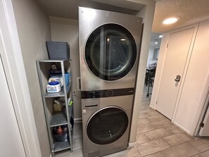 Une petite buanderie comprenant une machine à laver et un sèche-linge superposés, une étagère en métal où sont rangés les produits de lessive, et une porte donnant sur une autre pièce.