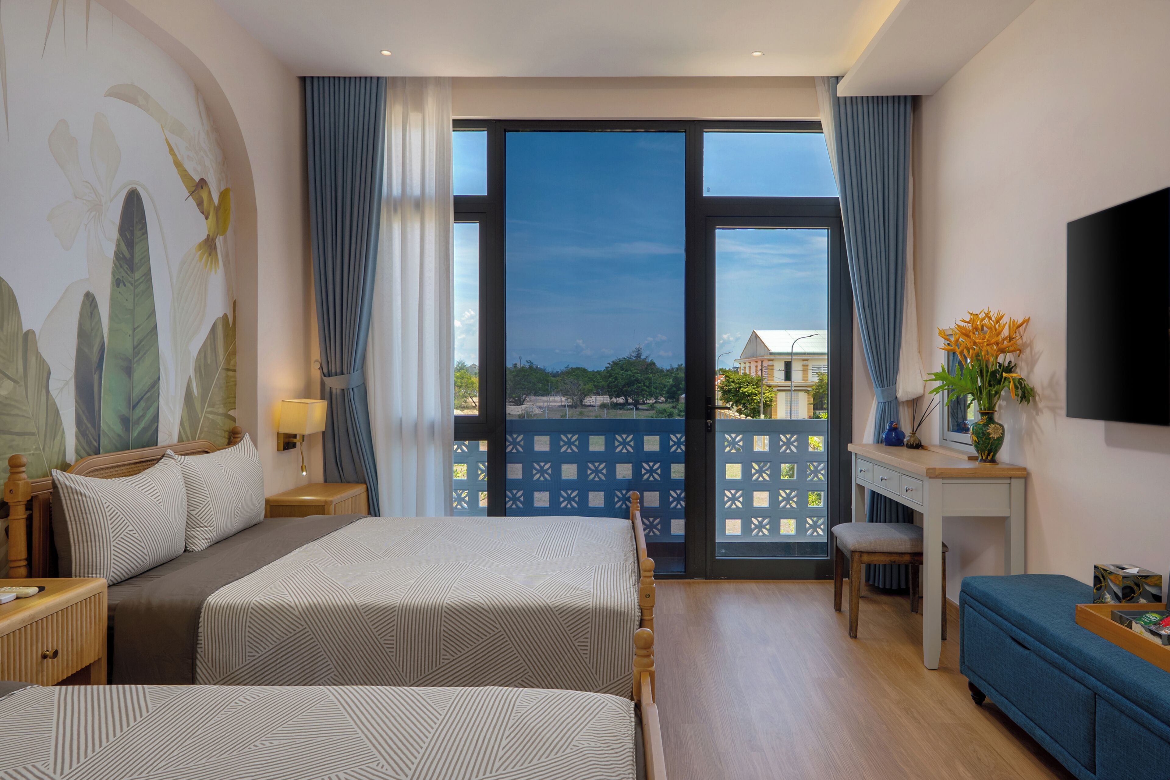 A hotel room with two beds, a desk, a TV, and a balcony with a view.