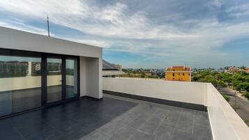 A rooftop terrace with a glass door leading to an indoor area, a flat roof with solar panels, and a view of surrounding buildings and greenery.