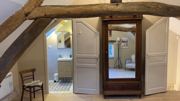 A room with a sloped ceiling, wooden beams, a wooden wardrobe, a chair, and a bathroom visible through an open door.