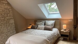 A bedroom with a sloped ceiling, a skylight, a bed with patterned bedding, a bedside table with a lamp, and a stone wall.