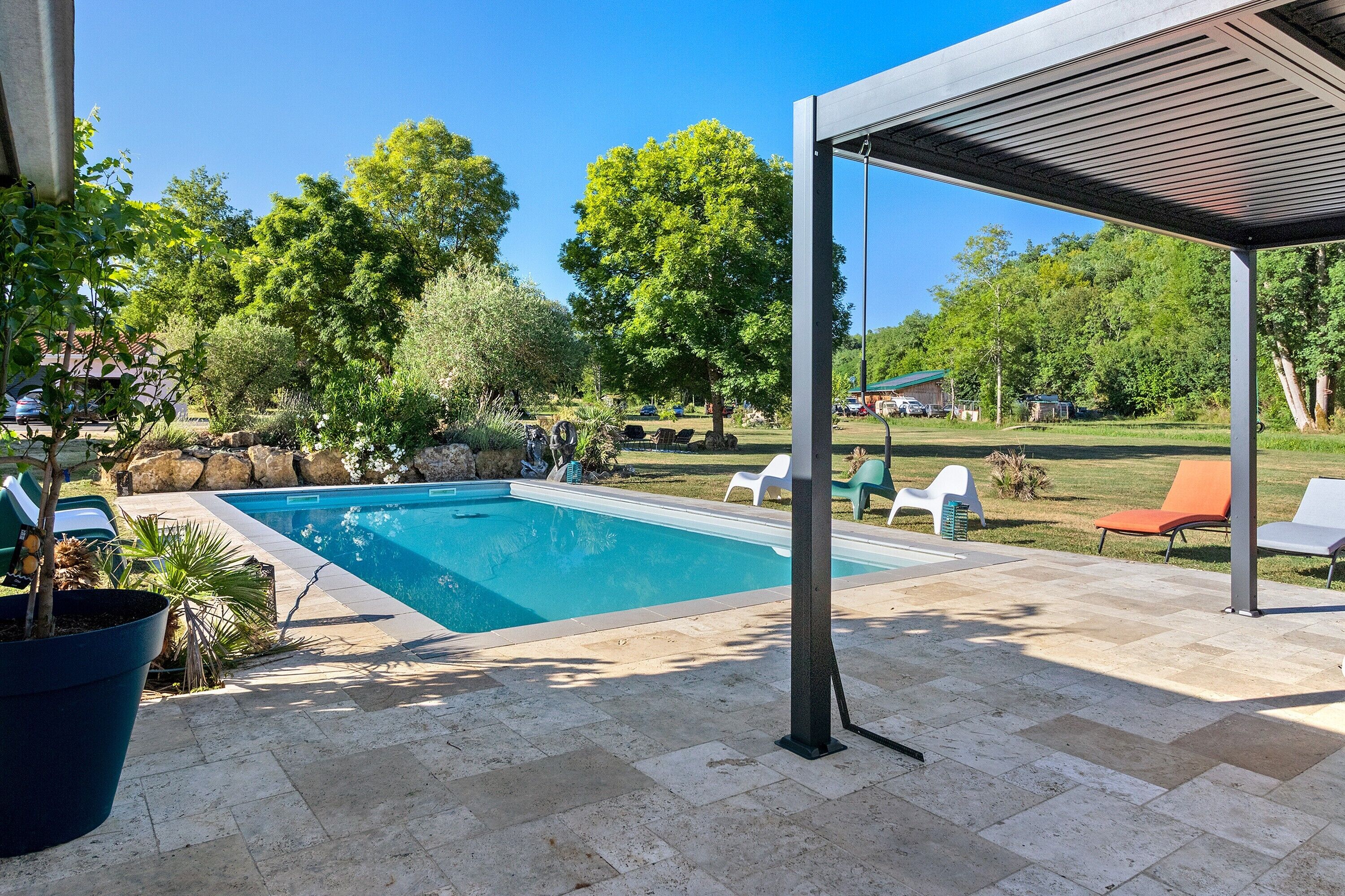 A swimming pool with a covered patio area, surrounded by greenery and outdoor furniture.
