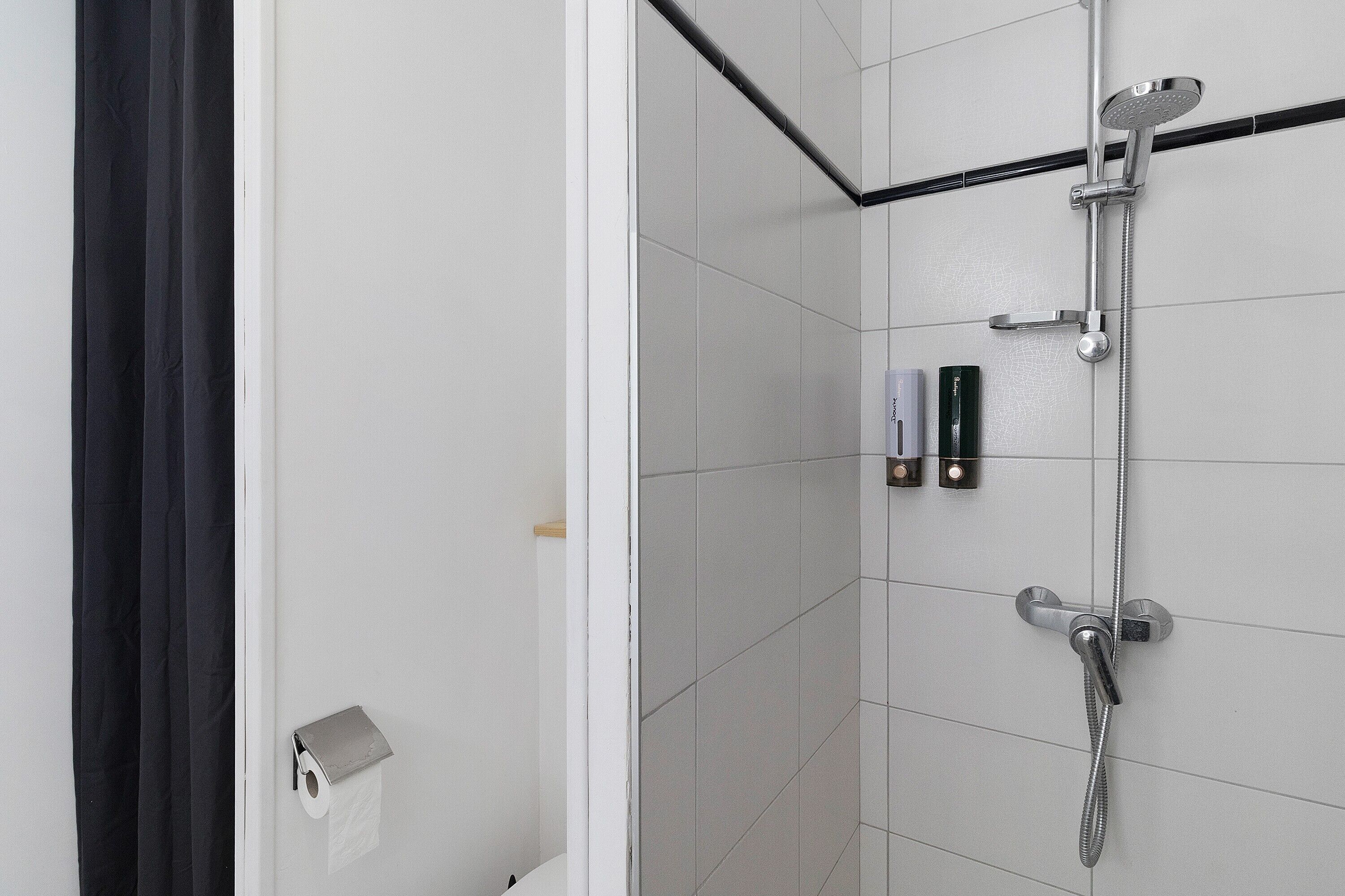 Bruser med håndbruser og vandhane på væggen, toiletpapirholder og sæbedispenser.