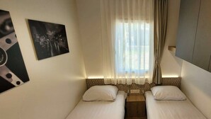 A small, well-lit room with two single beds, a window with sheer curtains, and a wall-mounted picture. - Holiday Home With 1 Bedroom (Berg en Terblijt)