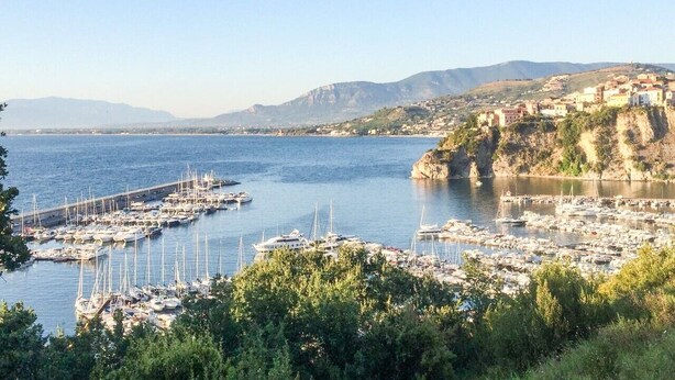 Un port marítim costaner amb vaixells, un moll i una població situada en un turó.