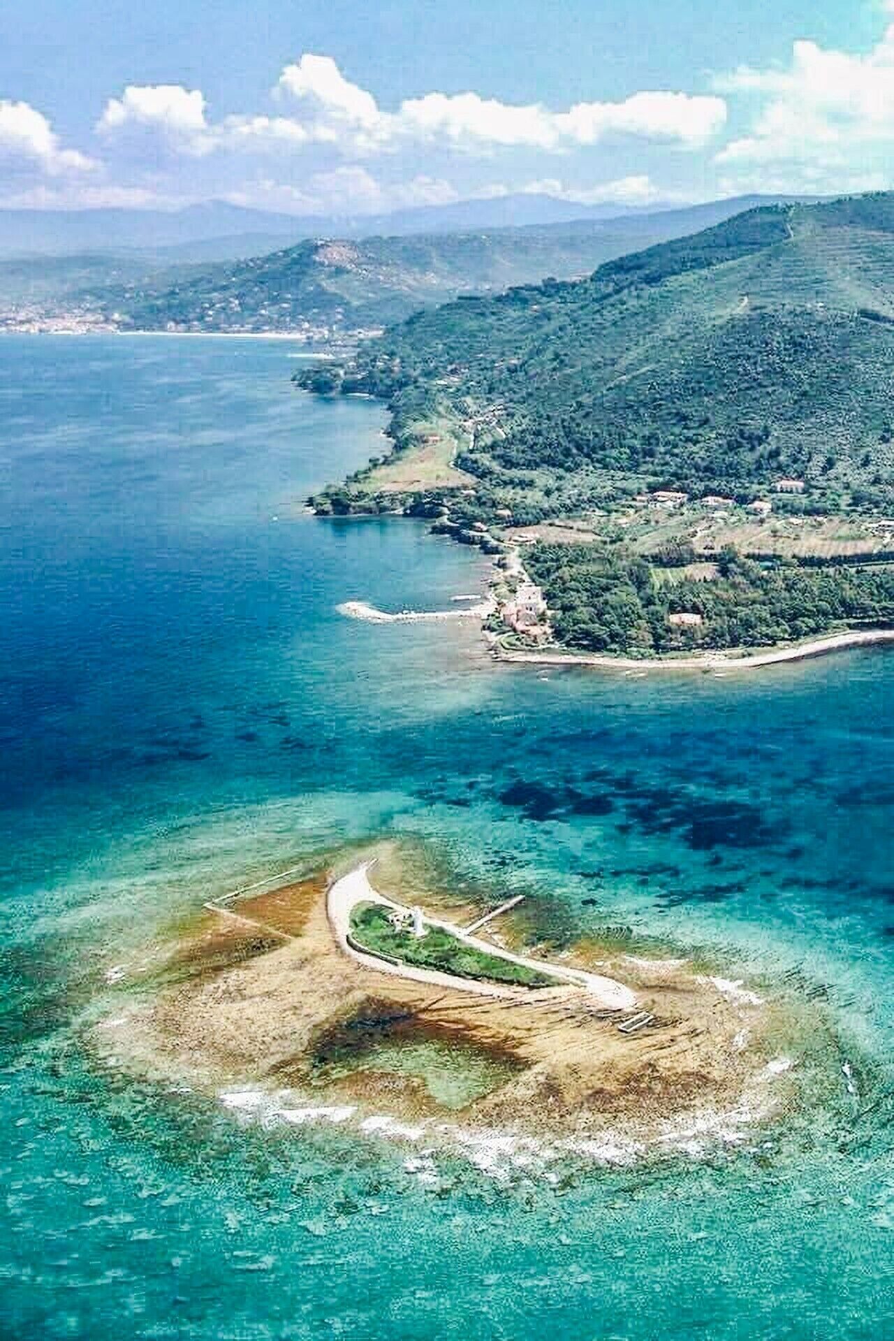 Une vue aérienne d’un paysage côtier, avec des eaux d’un bleu limpide, une petite île traversée par une route sinueuse et, au loin, une côte bordée de bâtiments.