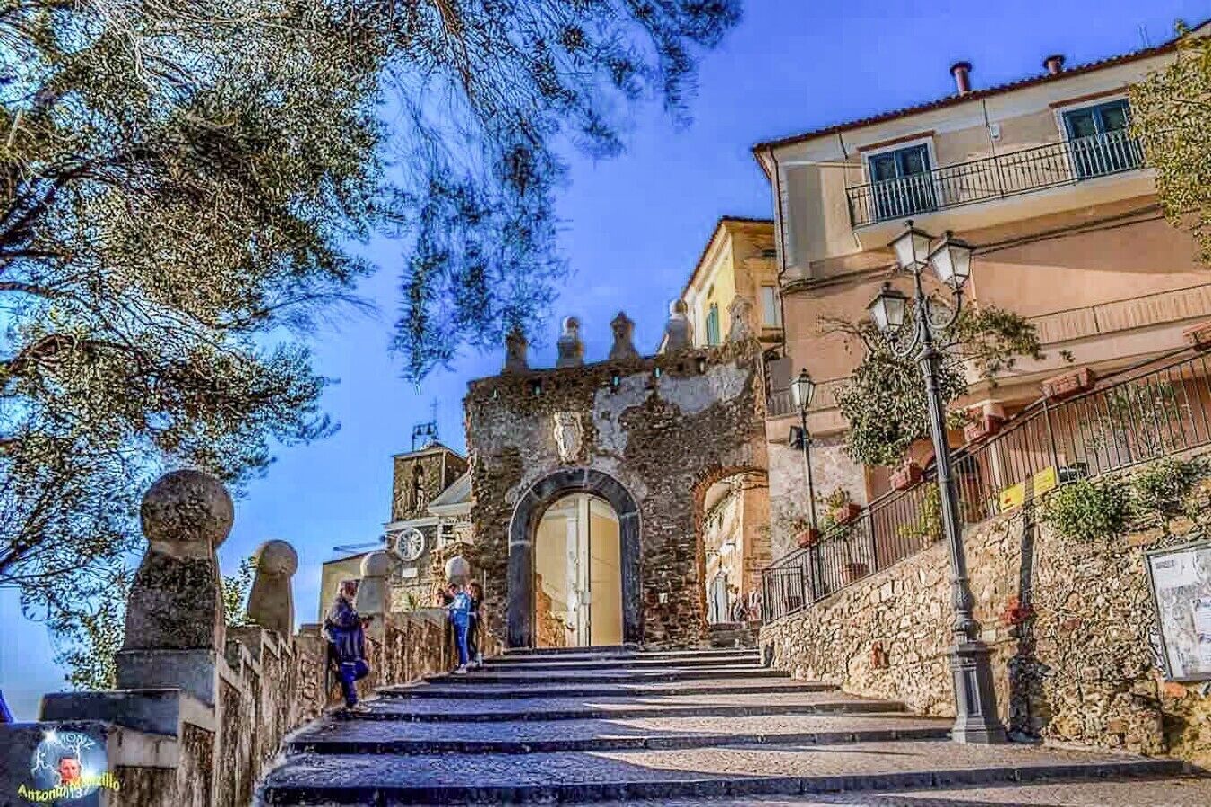 Una escalera de piedra conduce a un edificio histórico con un arco ornamentado, rodeado de árboles y edificios residenciales.