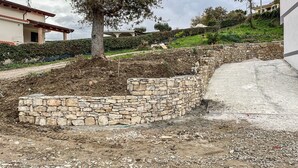 Un mur de pedra amb un disseny corbat, un camí de terra i un edifici al fons.