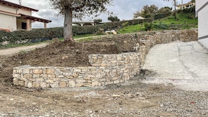 Een stenen keermuur met een gebogen ontwerp, een pad van aarde en een gebouw op de achtergrond.