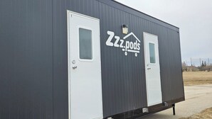 Une caravane grise avec deux portes blanches, un logo indiquant « ZZz.pods » sur le côté et une petite fenêtre sur la porte de gauche.