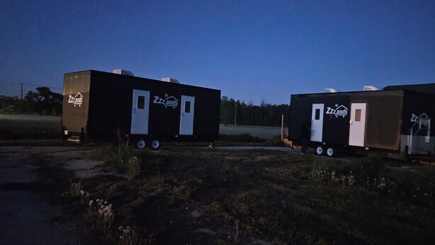 Deux unités mobiles arborant la marque « Zzz.pods », chacune dotée d’une porte et de fenêtres, sont stationnées sur un champ d’herbe.