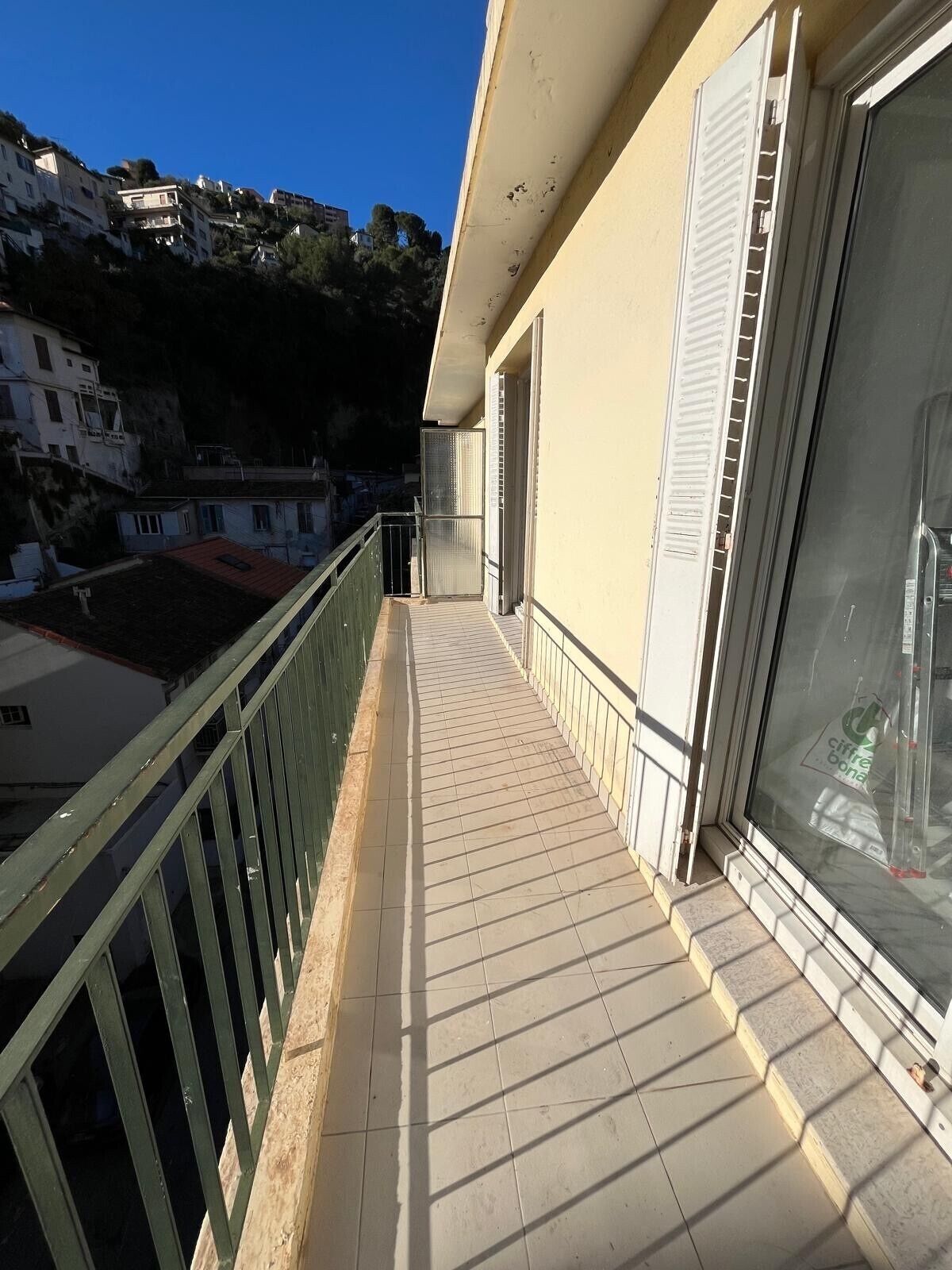 Un balcón con suelo de baldosas, baranda metálica y vistas a edificios al fondo.