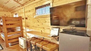 L’intérieur d’une cabane en bois comprend un coin cuisine, une table à manger avec des tabourets, un réfrigérateur et une fenêtre avec des rideaux.