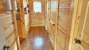 Un couloir intérieur aux murs en bois, avec une fenêtre au bout, des portes en bois et un pot en métal accroché au mur.