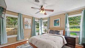 Ein Schlafzimmer mit einem Bett, einem Deckenventilator, einem Fenster mit Vorhängen, einem Heizkörper und einem Bild an der Wand.