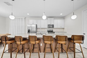 Cocina moderna con armarios blancos, refrigerador de acero inoxidable, microondas y horno empotrado. Hay taburetes de madera alrededor de una isla central con encimera de granito.