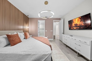 Quarto moderno com cama grande, televisão exibindo uma lareira, closet espaçoso e banheiro visível atrav és de uma porta aberta.