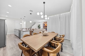 Uma cozinha moderna com armários brancos, uma sala de jantar com mesa e cadeiras de madeira e uma sala de estar com sofá e televisão.