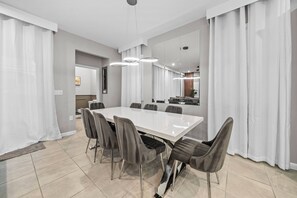 A modern dining area with a large rectangular table, grey upholstered chairs, and contemporary pendant lighting. - 6BR house with pool near Disney area (Kissimmee)