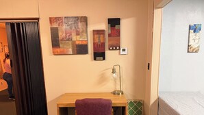 A room with a wooden desk, a purple chair, a bed with a white cover, a wall-mounted lamp, and a thermostat.
