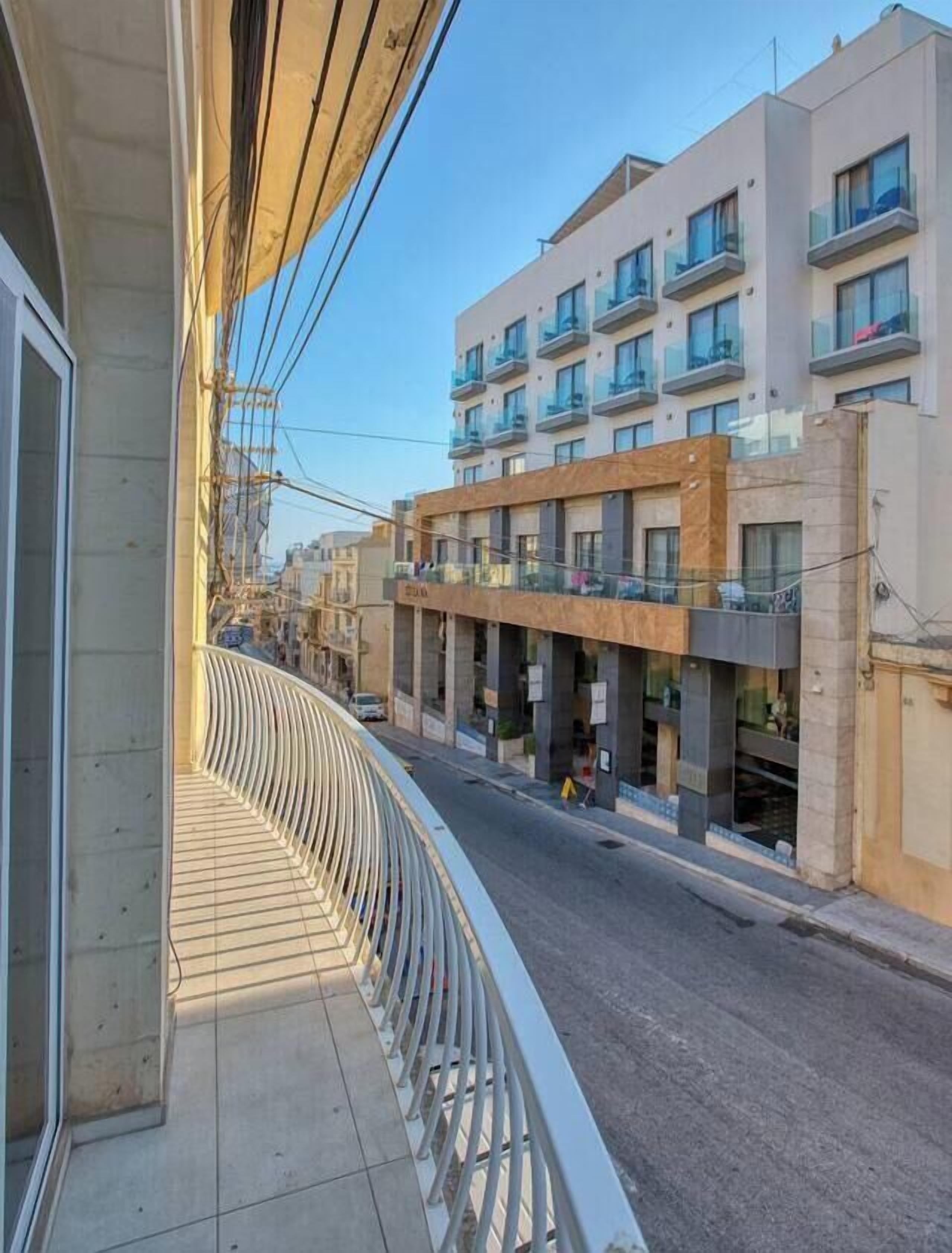 Un balcon doté d’une balustrade incurvée, donnant sur une rue bordée d’immeubles, dont un immeuble résidentiel à plusieurs étages également pourvu de balcons.