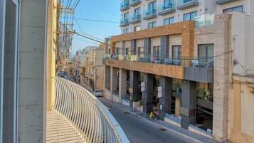 Ein Balkon mit geschwungener Geländer, der auf eine Straße mit Gebäuden blickt, darunter ein mehrstöckiges Wohngebäude mit Balkonen.