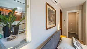A hotel room with a bed, a bedside table with a plant, a framed picture on the wall, and a hallway leading to another room. - Alba Palace Luxury Apartments - White (Alba)