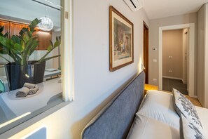 Ein Hotelzimmer mit Bett, Nachttisch mit Pflanze, gerahmtes Bild an der Wand und einem zur anderen Kammer führenden Flur.