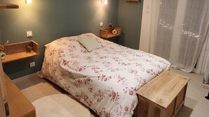 Une chambre à coucher comprenant un lit, des tables de chevet, un banc en bois, une étagère fixée au mur et une fenêtre avec des rideaux.