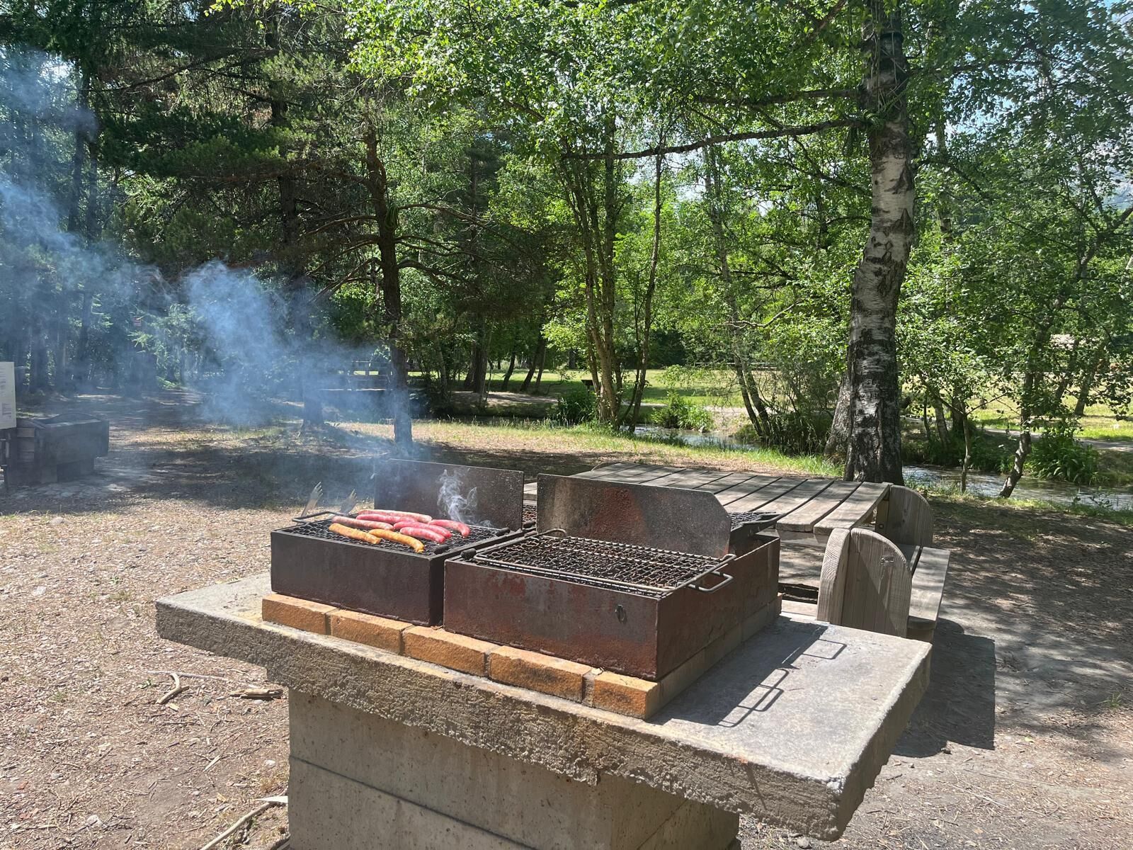 A barbecue grill with sausages and a grill cover, surrounded by trees and a picnic table.