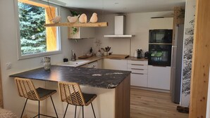 Une cuisine moderne dotée d’un îlot central, de chaises en bois, d’un four encastré et d’une fenêtre donnant sur les arbres.
