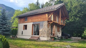 Une cabane en bois à deux étages, avec des fondations en pierre, un balcon et une voiture garée à l’extérieur.