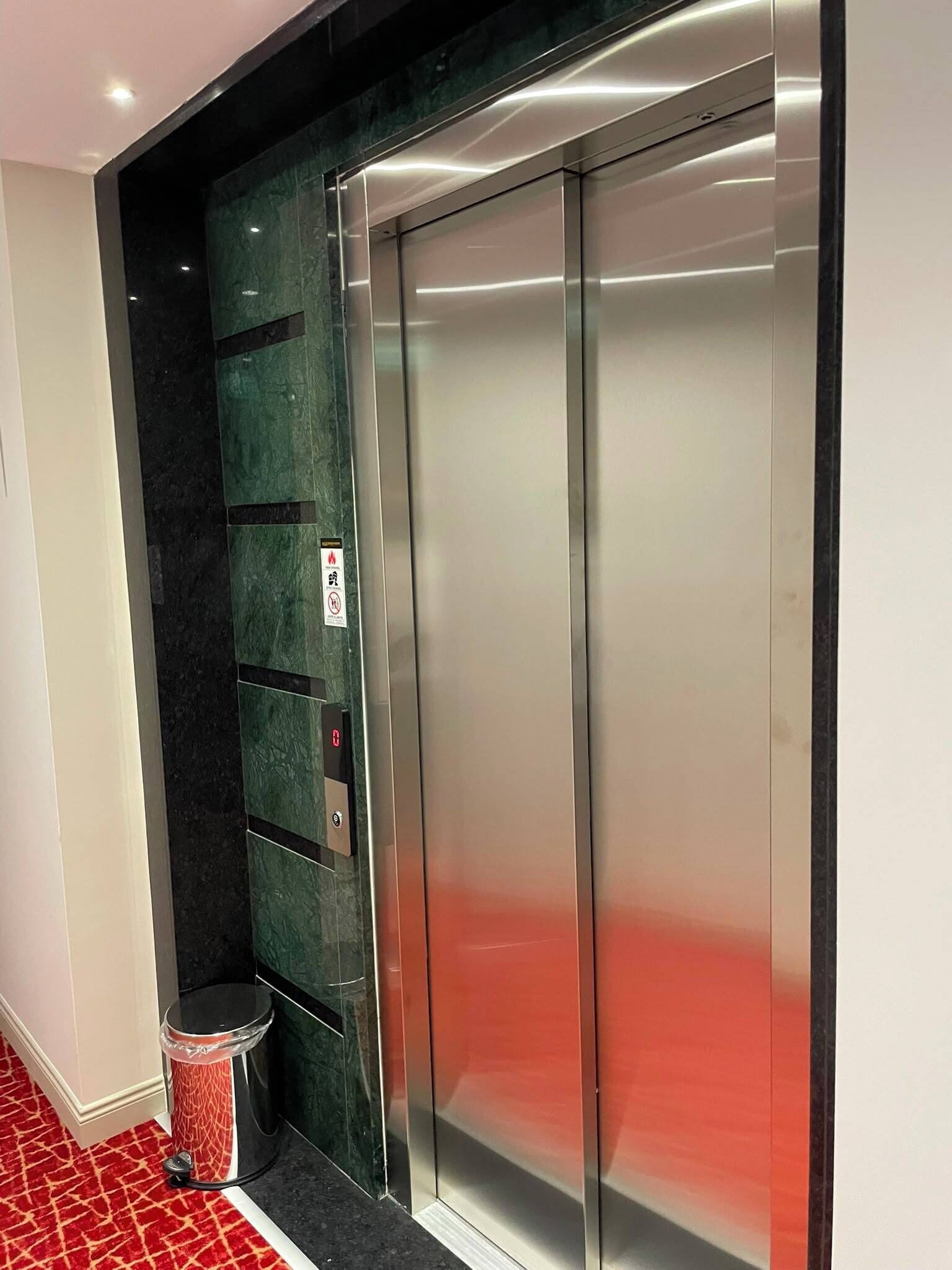 An elevator with a metallic finish, a fire alarm button, and a trash bin.