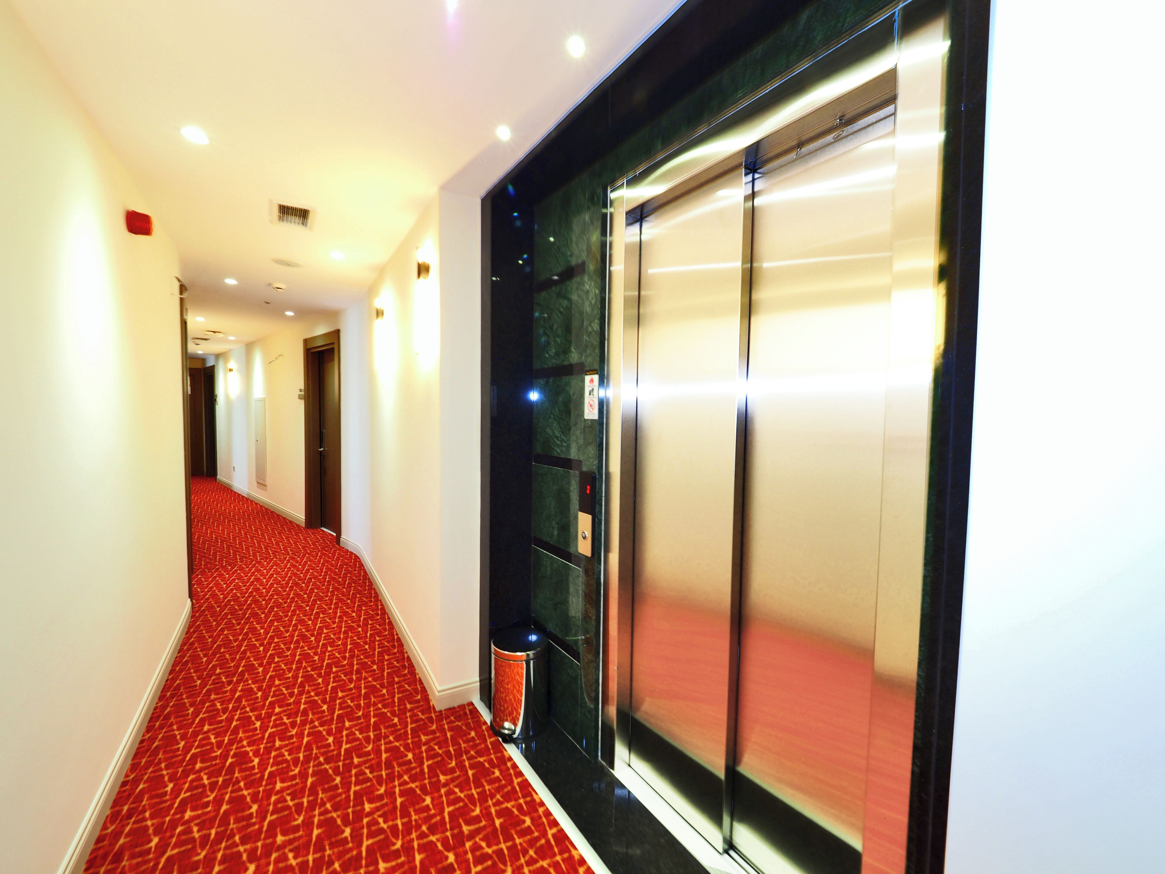 Un couloir d’hôtel avec un tapis rouge, un ascenseur en verre et une porte au bout.