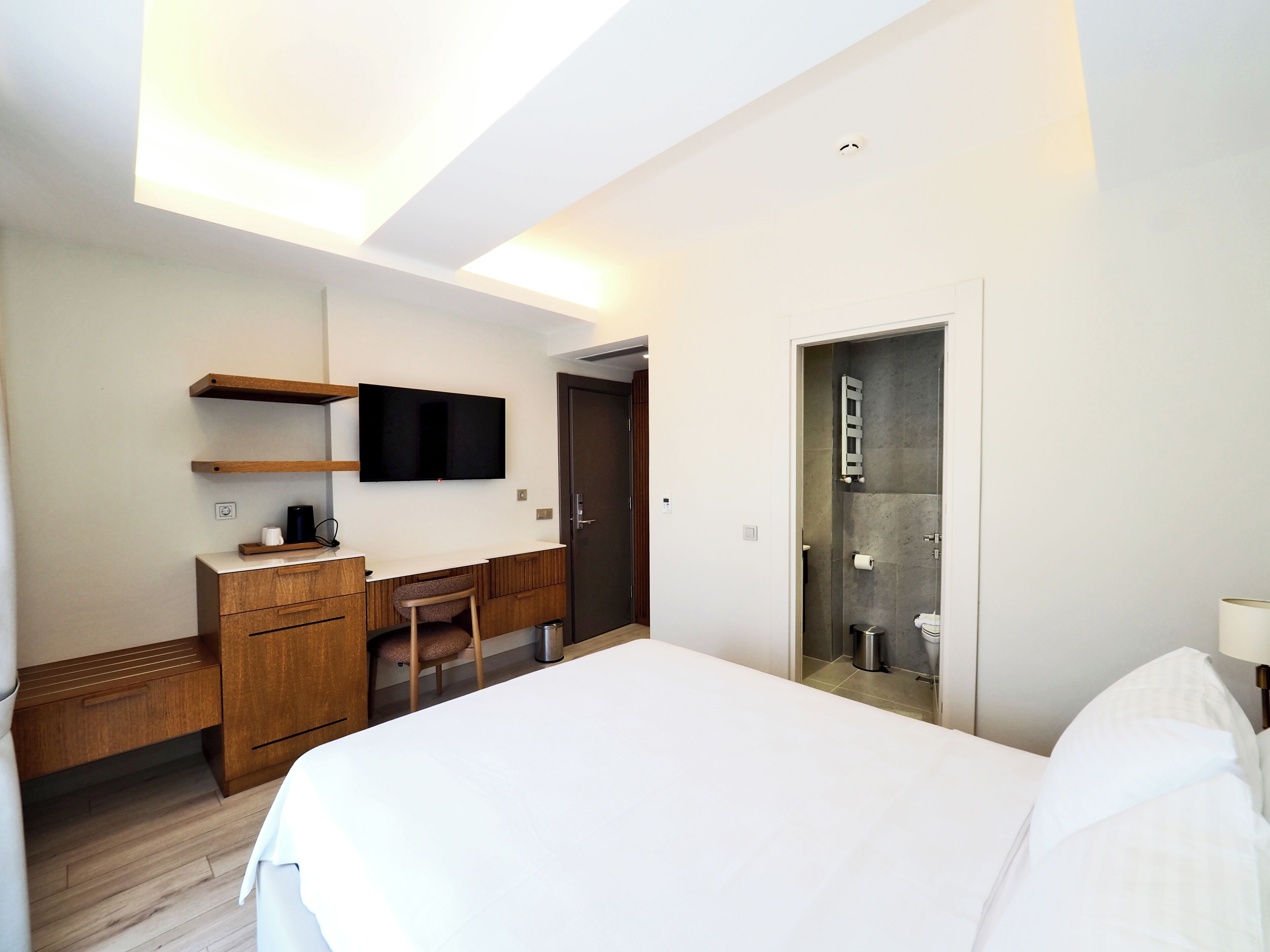 Quarto de hotel com uma cama, uma escrivaninha com cadeira, televisão fixada na parede, chuveiro e uma porta.