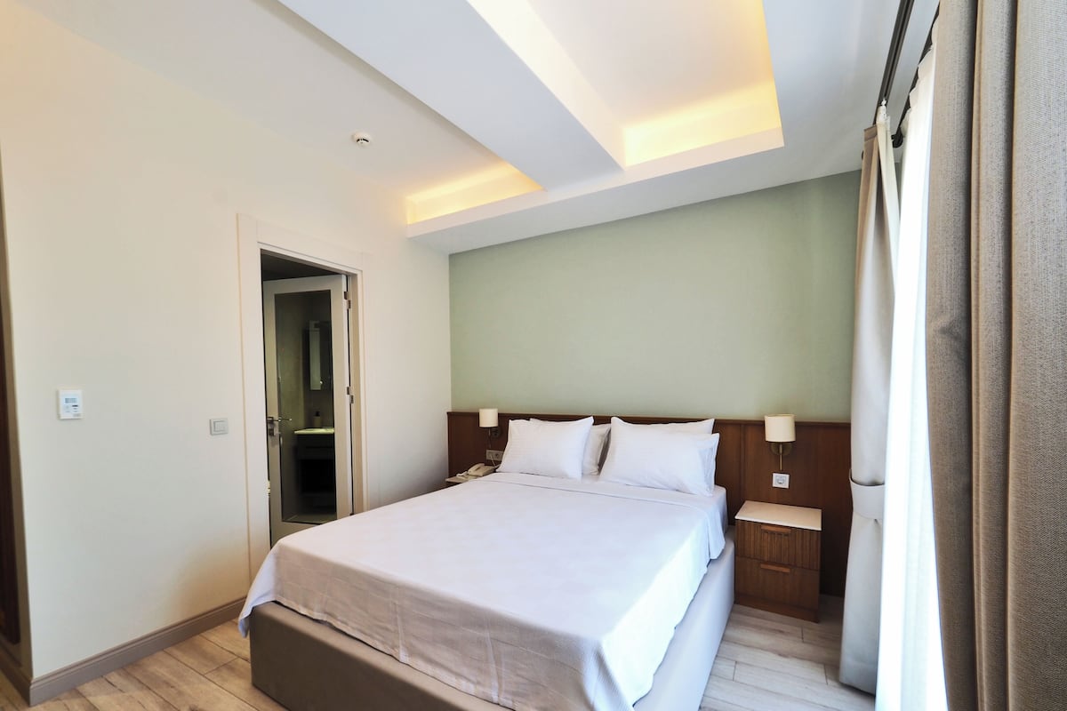 A hotel room with a bed, bedside tables, a nightstand, a door to a bathroom, and a light fixture on the ceiling.