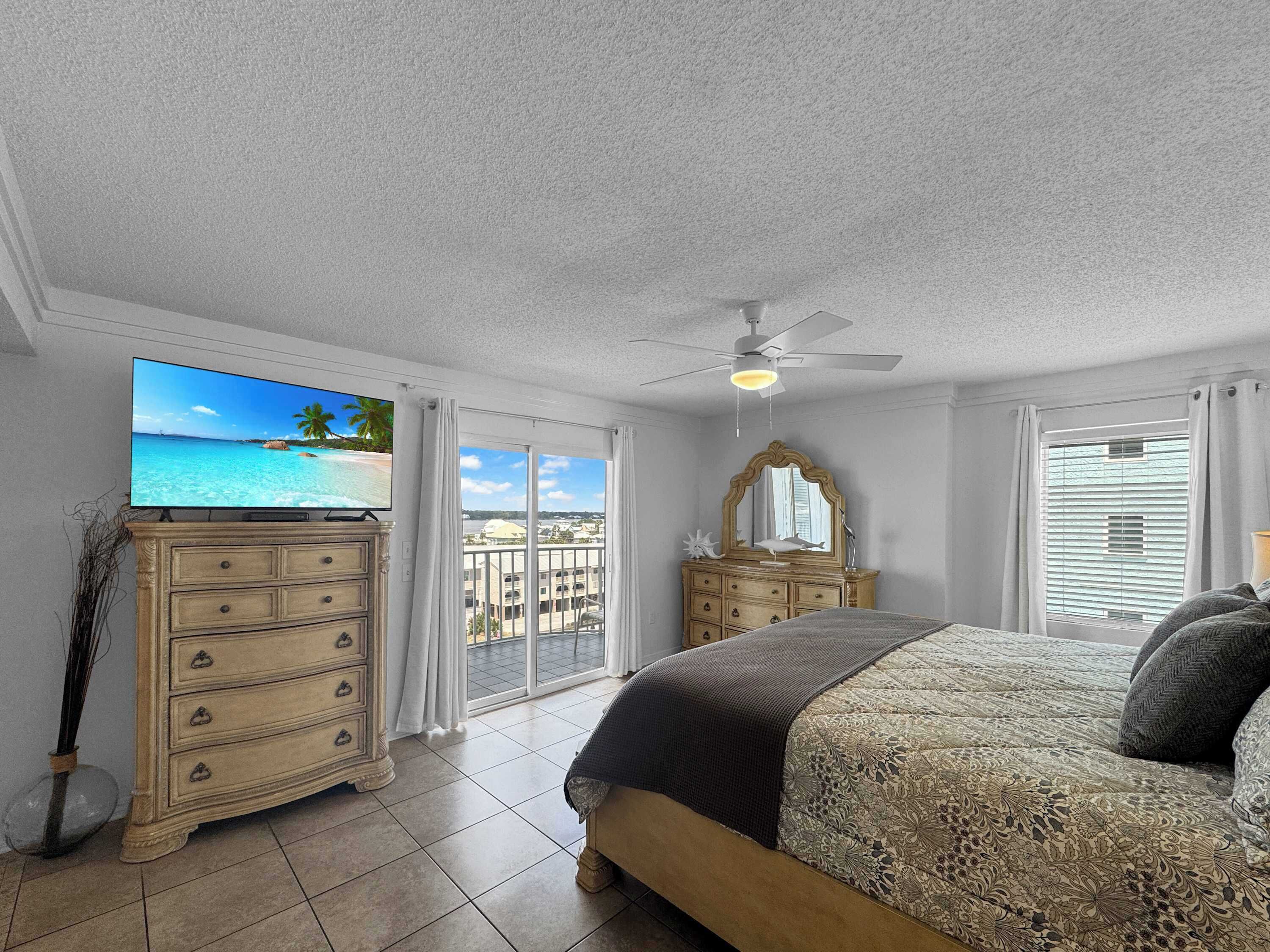 A bedroom with a bed, a dresser, a television, a ceiling fan, and a view of the ocean through large windows.