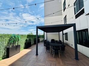 Un patio al aire libre con una pérgola, asientos e iluminación con luces colgantes, frente a un edificio moderno.