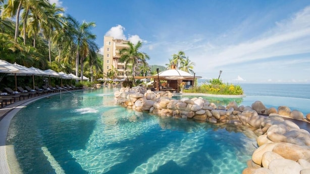 Una piscina di resort con acqua cristallina, circondata da palme e un edificio con tetto a cupola.