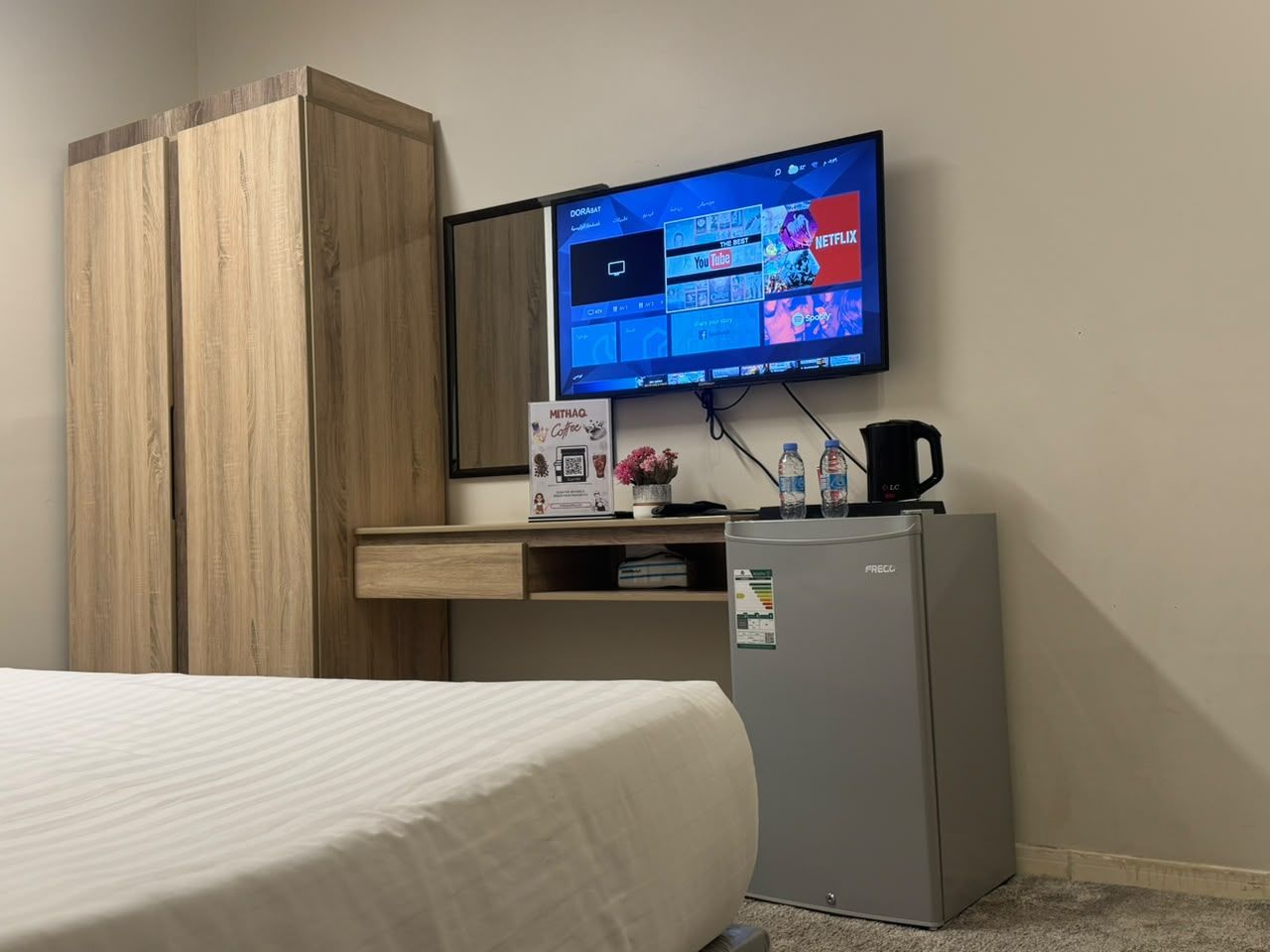 Camera d'albergo con un letto, una scrivania con un monitor che mostra servizi di streaming, un frigorifero e una macchina per il caffè.